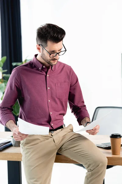 Attentive businessman holding documents while sitting on desk near coffee to go — Stock Photo