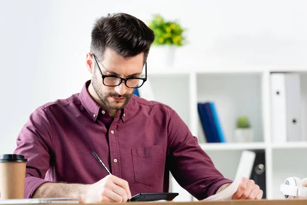 Selektiver Fokus des aufmerksamen Geschäftsmannes, der Papier hält und in der Nähe von Taschenrechner und Coffee to go schreibt — Stockfoto