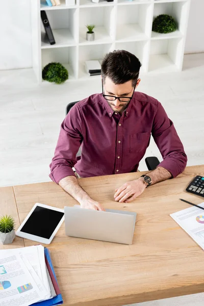 Vista de ángulo alto del hombre de negocios serio usando el ordenador portátil cerca de la tableta digital con la pantalla en blanco y documentos - foto de stock