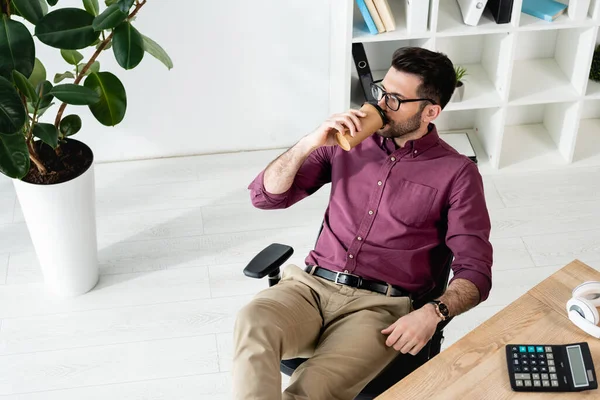 Vista de ángulo alto de joven empresario beber café para ir sentado en el lugar de trabajo cerca de la planta en maceta - foto de stock