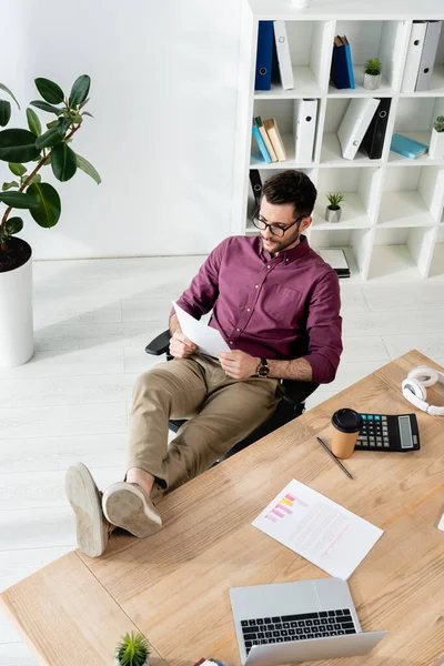 Junger Geschäftsmann liest Dokument, während er mit Beinen auf dem Schreibtisch neben Laptop, Taschenrechner und Coffee to go sitzt — Stockfoto