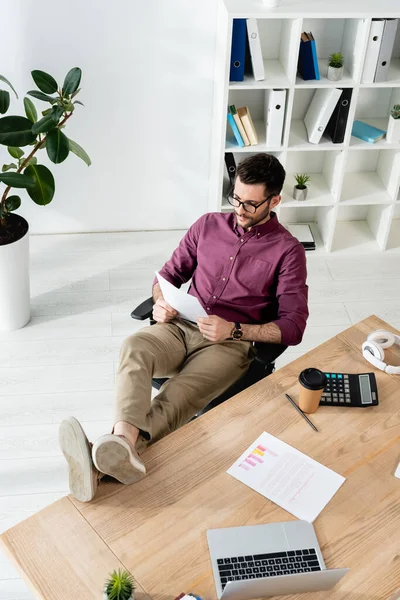 Joven hombre de negocios leyendo documento mientras está sentado con las piernas en el escritorio cerca de la computadora portátil, calculadora y café para llevar - foto de stock