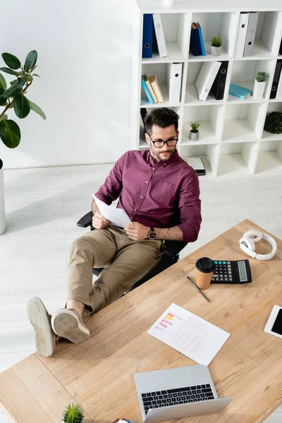 Vue grand angle de l'homme d'affaires tenant le document tout en étant assis avec les jambes sur le bureau près de l'ordinateur portable, calculatrice et café à emporter — Photo de stock