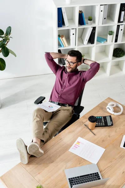 High angle of businessman sitting with legs on desk and hands behind head near gadgets, papers and disposable cup — Stock Photo