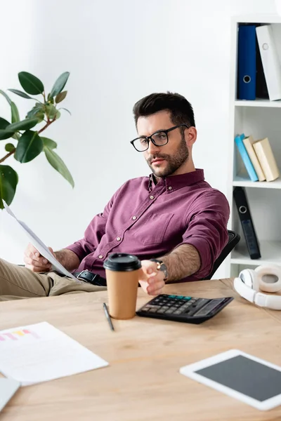 Hombre de negocios guapo sentado en el lugar de trabajo, sosteniendo el documento y tomando café para ir — Stock Photo