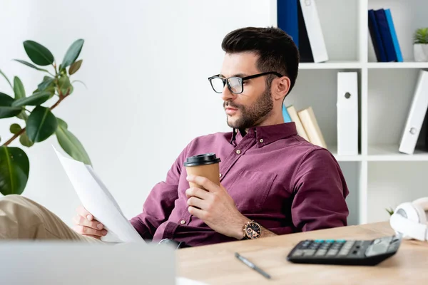 Вибірковий фокус молодого бізнесмена, який тримає паперовий стаканчик під час читання документа на робочому місці — стокове фото