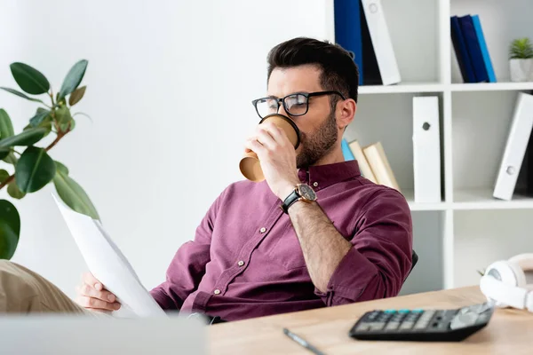 Вибірковий фокус молодого бізнесмена, який п'є каву, щоб піти під час вивчення документа на робочому місці — стокове фото