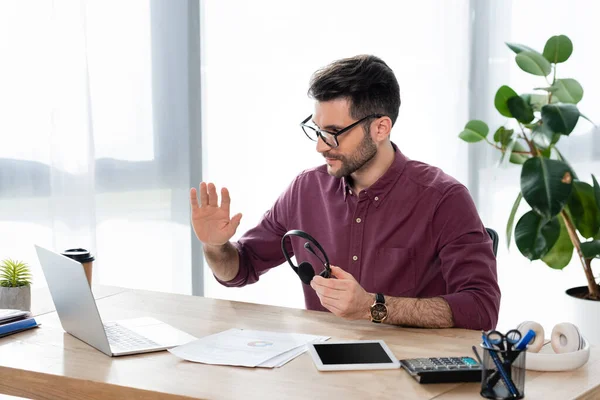 Giovane uomo d'affari che tiene le cuffie e agita la mano durante la videochiamata sul laptop — Foto stock