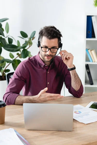 Joven hombre de negocios en auriculares hablando mientras tiene videollamada en el ordenador portátil - foto de stock