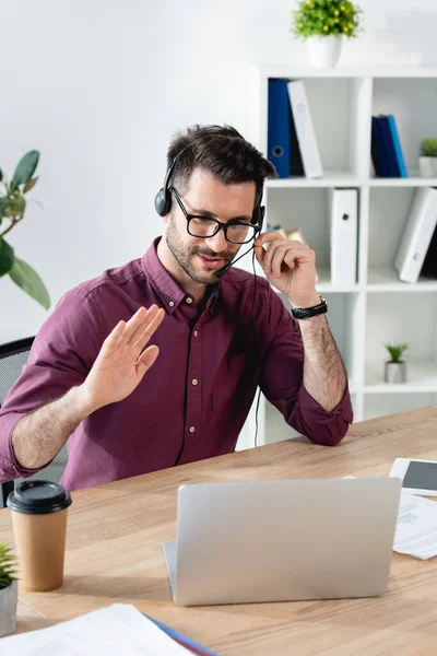 Homem de negócios sorridente no fone de ouvido mostrando gesto de Olá durante o bate-papo por vídeo no laptop — Fotografia de Stock