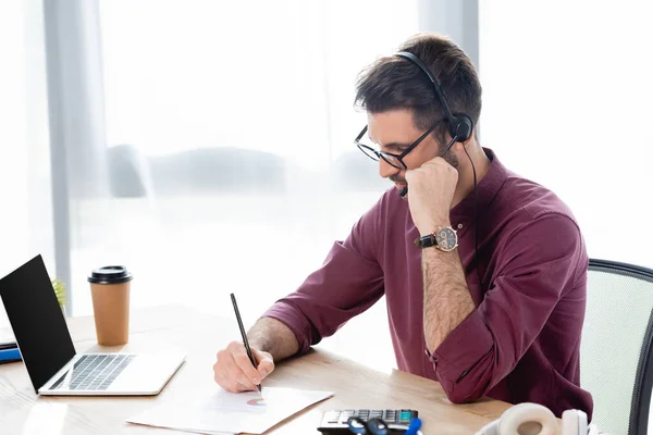 Empresario concentrado en la escritura de auriculares en papel durante la conferencia en línea en el ordenador portátil - foto de stock