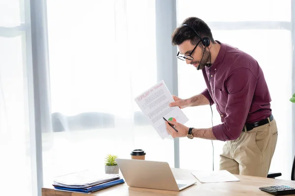 Jovem empresário em fone de ouvido apontando com lápis no documento durante reunião on-line no laptop — Fotografia de Stock