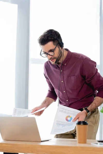 Giovane uomo d'affari guardando laptop e in possesso di documenti con infografica durante la videochiamata — Foto stock