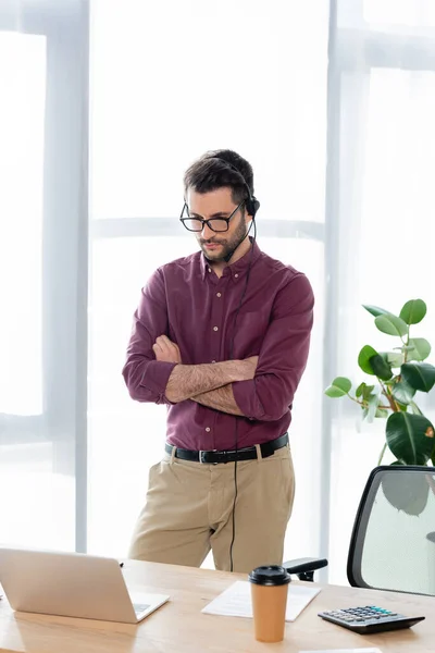 Jeune homme d'affaires sérieux dans un casque debout avec les bras croisés pendant le chat vidéo sur ordinateur portable — Photo de stock