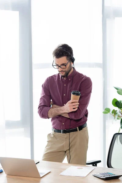 Young, serious businessman in headset holding coffee to go during online meeting on laptop — Stock Photo