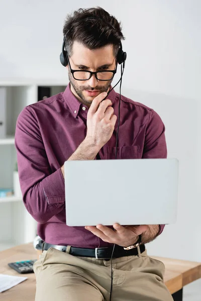 Jeune homme d'affaires sérieux dans un casque tenant un ordinateur portable pendant le chat vidéo — Photo de stock