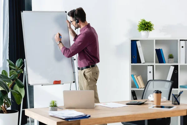 Young businessman in headset writing on clipboard near desk with laptop — Stock Photo