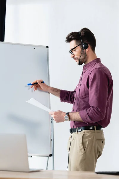 Konzentriert Geschäftsmann in Headset-Schrift auf Flipchart, während Dokument in der Nähe Schreibtisch mit Laptop — Stockfoto
