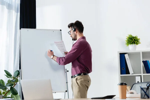 Young businessman in headset writing on flipchart while holding document near desk with laptop — Stock Photo