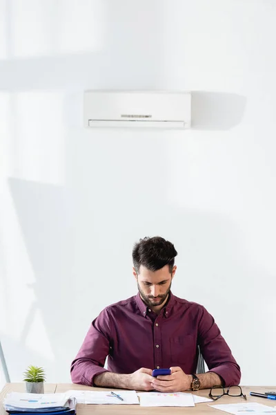 Joven hombre de negocios usando teléfono inteligente mientras está sentado en el lugar de trabajo bajo el aire acondicionado - foto de stock