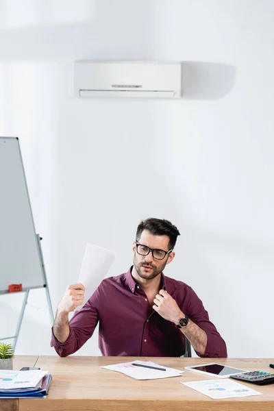 Joven hombre de negocios tocando camisa y agitando papel mientras sufre de calor en la oficina - foto de stock