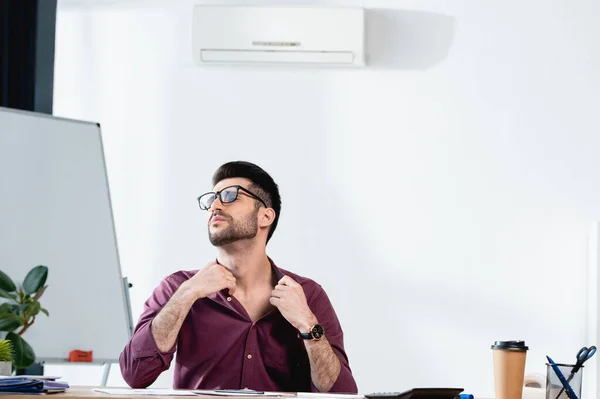 Exhausted businessman sitting at workplace and touching shirt while suffering from heat in office — Stock Photo