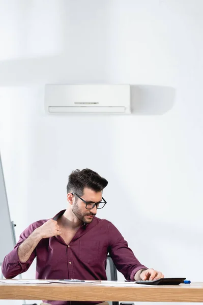 Junger Geschäftsmann sitzt am Arbeitsplatz und berührt Hemd, während er im Büro unter Hitze leidet — Stockfoto