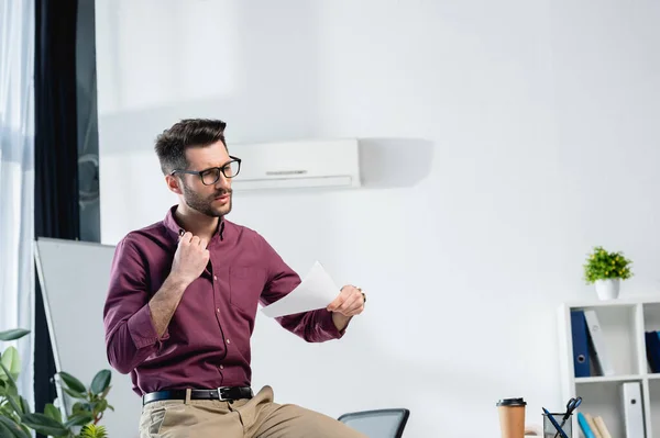 Erschöpfter Geschäftsmann sitzt am Schreibtisch, berührt Hemd und wedelt mit Papier, während er im Büro unter Hitze leidet — Stockfoto