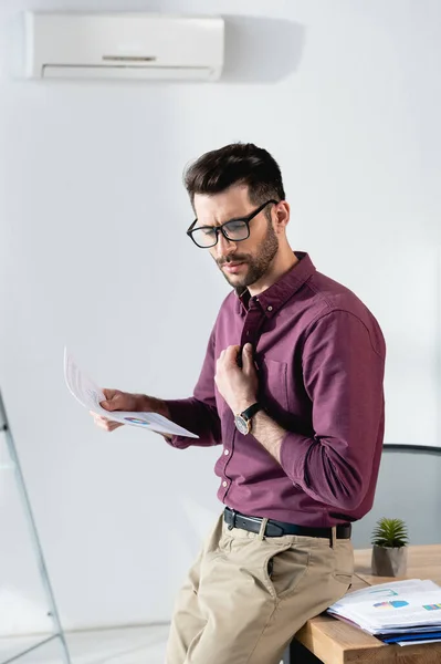 Displeased businessman holding paper and touching shirt while suffering from heat in office — Stock Photo
