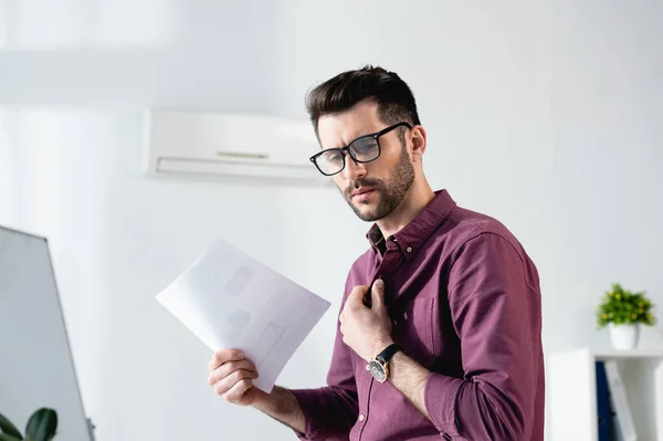 Displeased businessman waving paper and touching shirt while suffering from heat in office — Stock Photo