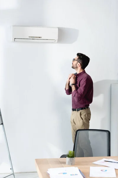 Молодий бізнесмен торкається сорочки, стоячи під кондиціонером і страждаючи від спеки — стокове фото