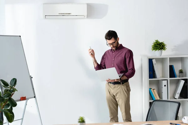 Schöner Geschäftsmann mit Laptop und eingeschalteter Klimaanlage mit Fernbedienung — Stockfoto