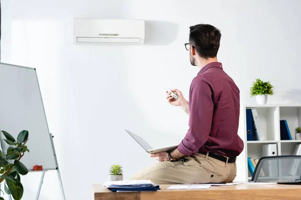 Uomo d'affari seduto sulla scrivania con computer portatile e accendere condizionatore d'aria con telecomando — Foto stock