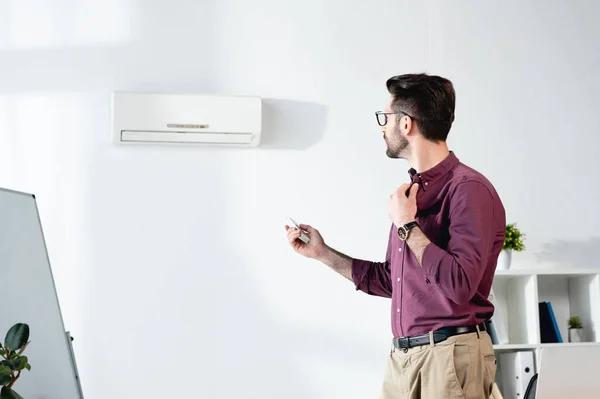 Jeune homme d'affaires souffrant de chaleur, chemise touchante et allumage du climatiseur avec télécommande — Photo de stock