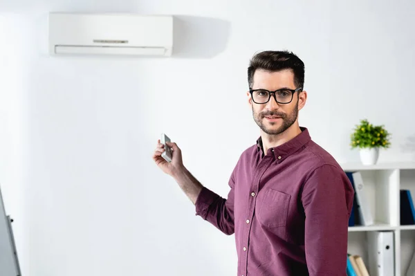 Feliz hombre de negocios sonriendo a la cámara mientras enciende el aire acondicionado con mando a distancia - foto de stock