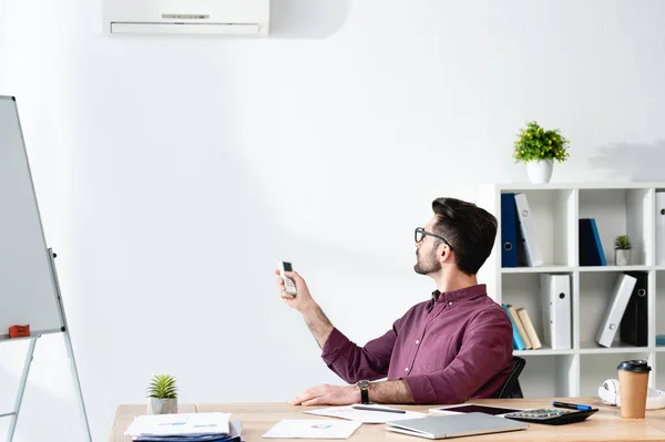 Joven empresario sentado en el lugar de trabajo y encendiendo el aire acondicionado con mando a distancia - foto de stock