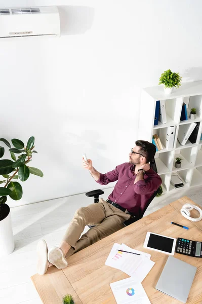 Vista de ángulo alto del hombre de negocios sentado con las piernas en la mesa y encender el aire acondicionado con mando a distancia - foto de stock