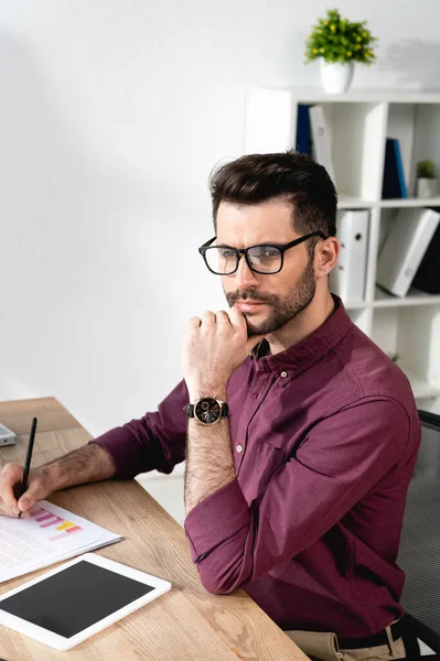 Thoughtful businessman writing on paper with charts near digital tablet with blank screen — Stock Photo