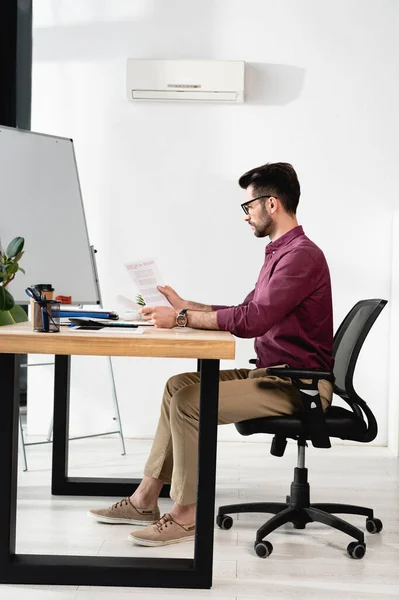 Side view of businessman looking at papers while sitting at workplace near air conditioner — Stock Photo