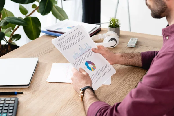 Vista recortada del empresario sosteniendo papel con analítica cerca de la computadora portátil y auriculares inalámbricos - foto de stock