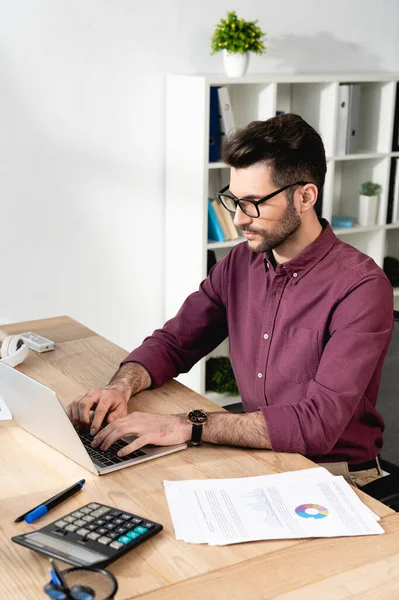 Homme d'affaires concentré tapant sur ordinateur portable près de documents et calculatrice — Photo de stock