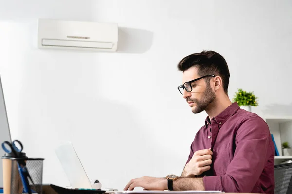 Junger Geschäftsmann berührt Hemd während der Arbeit am Laptop und leidet unter Hitze in der Nähe von Klimaanlage — Stockfoto