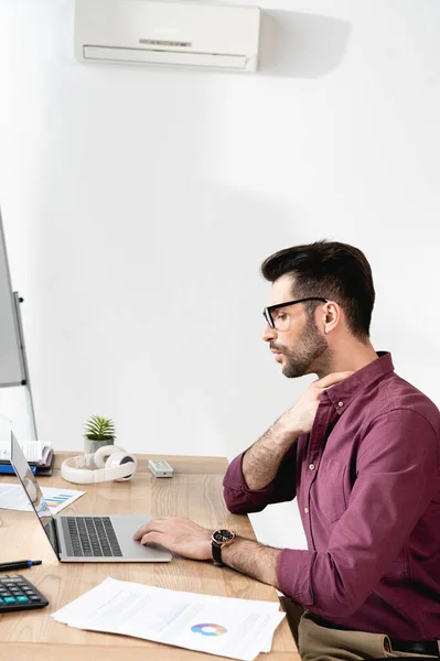Jeune homme d'affaires travaillant sur ordinateur portable et touchant le cou tout en souffrant de chaleur sous climatiseur — Photo de stock