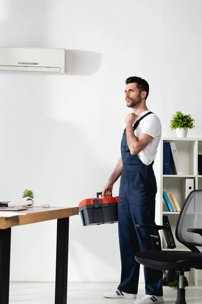 Bellissimo riparatore in piedi con cassetta degli attrezzi vicino al condizionatore d'aria rotto e affetto da calore — Foto stock