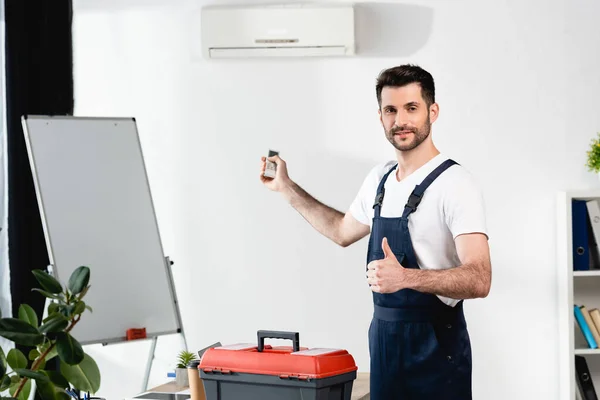 Smiling worker holding remote controller and showing thumb up while standing near toolbox and air conditioner — Stock Photo