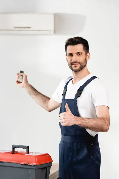 Réparateur souriant tenant télécommande et montrant pouce près de la boîte à outils et climatiseur sur le mur — Photo de stock