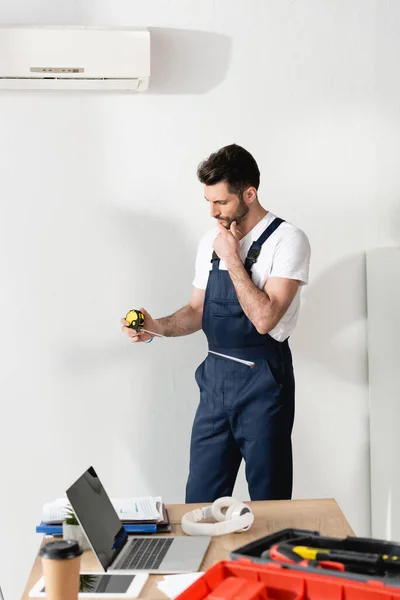 Thoughtful repairman holding measuring tape while standing under broken air conditioner — Stock Photo