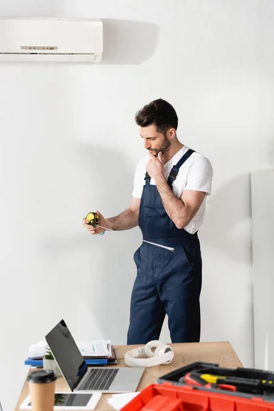 Nachdenklicher Reparateur hält Maßband in der Hand, während er unter defekter Klimaanlage steht — Stockfoto