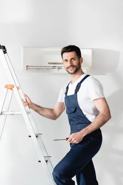 Smiling repairman holding screwdriver while standing on stepladder near air conditioner — Stock Photo