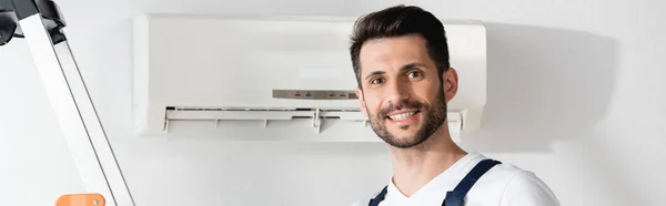 Raccolto panoramico di riparatore felice sorridente alla macchina fotografica vicino al condizionatore d'aria sulla parete — Foto stock